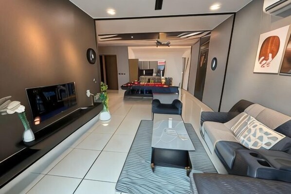 A modern living room with a flat-screen TV, a sofa, a coffee table, and a dining area in the background. - Green Haven Three-Bedroom (Plentong)