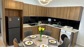 Een kleine keuken met een inbouwoven, een magnetron en een spoelbak. Er is een eettafel met stoelen en een vaas met bloemen.