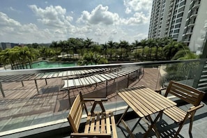 A rooftop terrace with a pool, wooden furniture, and a view of palm trees and buildings.