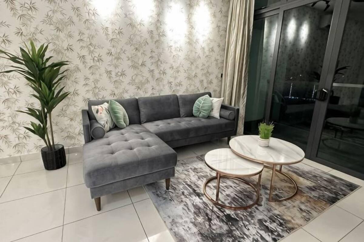 A living room with a grey sectional sofa, two round coffee tables, a potted plant, and a large window with curtains.