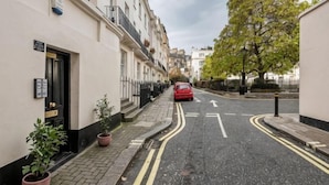 A narrow street with terraced houses, a red car parked on the side, and a tree on the right. - Kula London - Victoria (London)