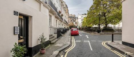 A narrow street with terraced houses, a red car parked on the side, and a tree on the right.