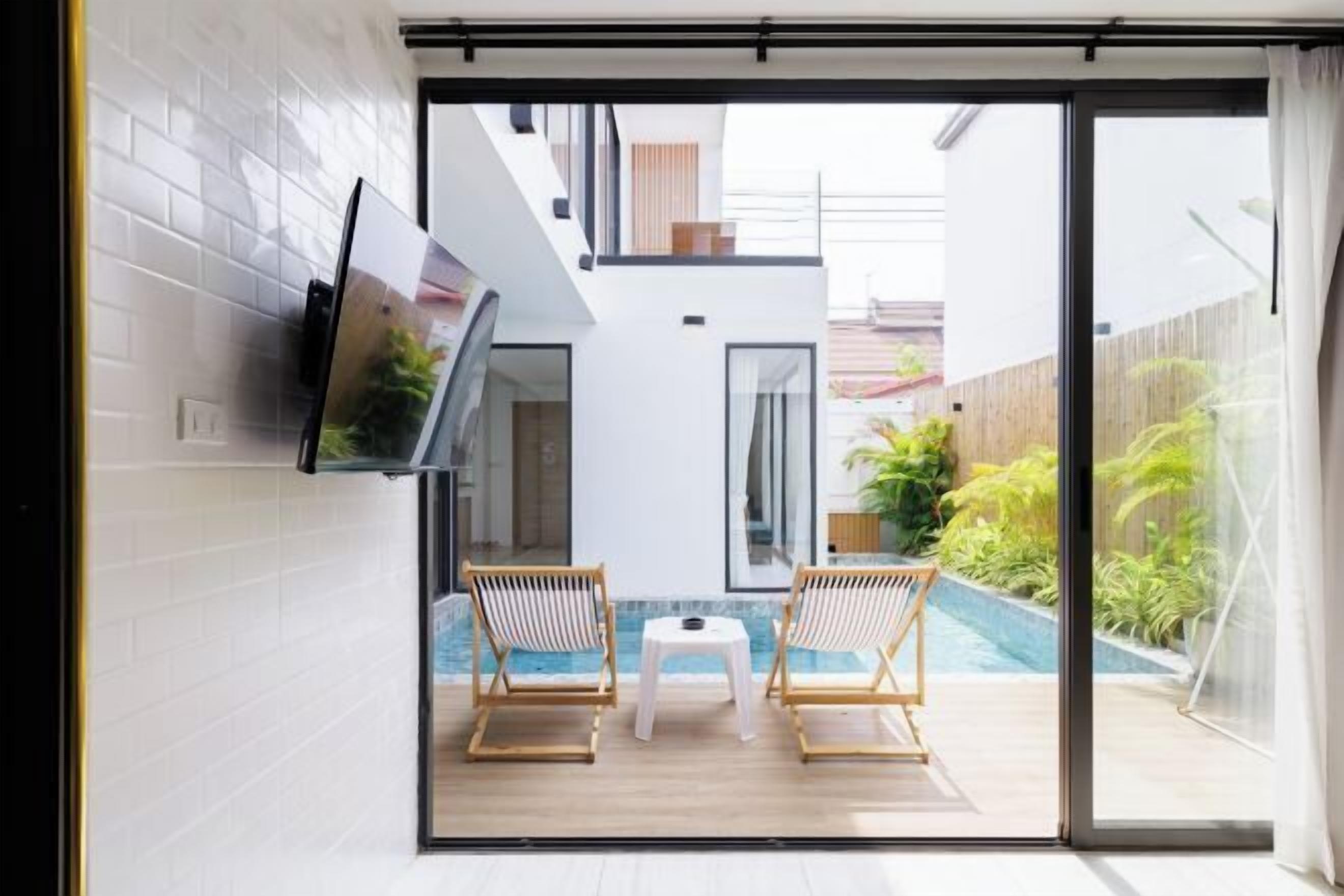 Un intérieur moderne avec un téléviseur fixé au mur, un espace piscine agrémenté de deux chaises en bois et d’une petite table, et une porte-fenêtre coulissante donnant sur un espace extérieur.