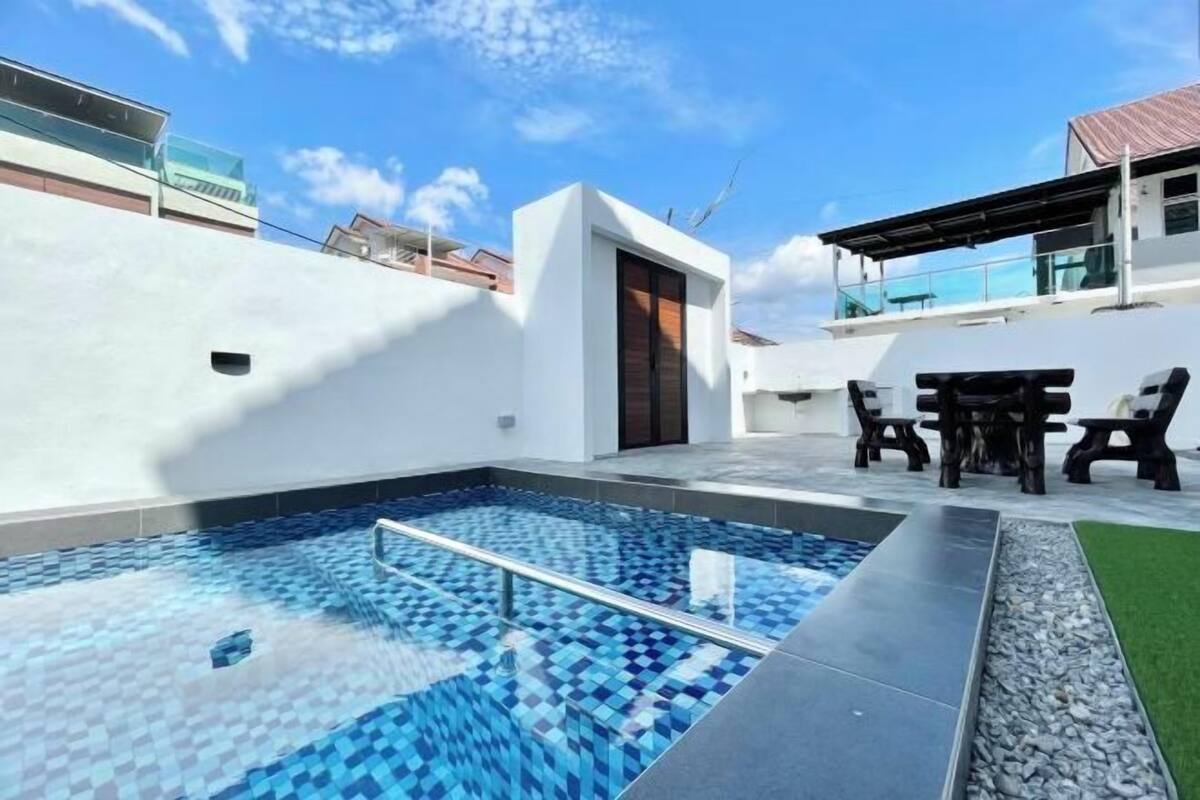 A swimming pool with a tiled deck, a small outdoor seating area, and a white building with a door.