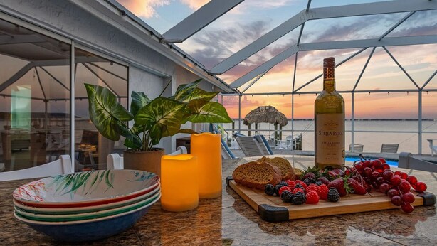A table with plates, a candle, a bottle of Stella Rosa wine, and a fruit platter on a balcony with a view of the sunset and beach.