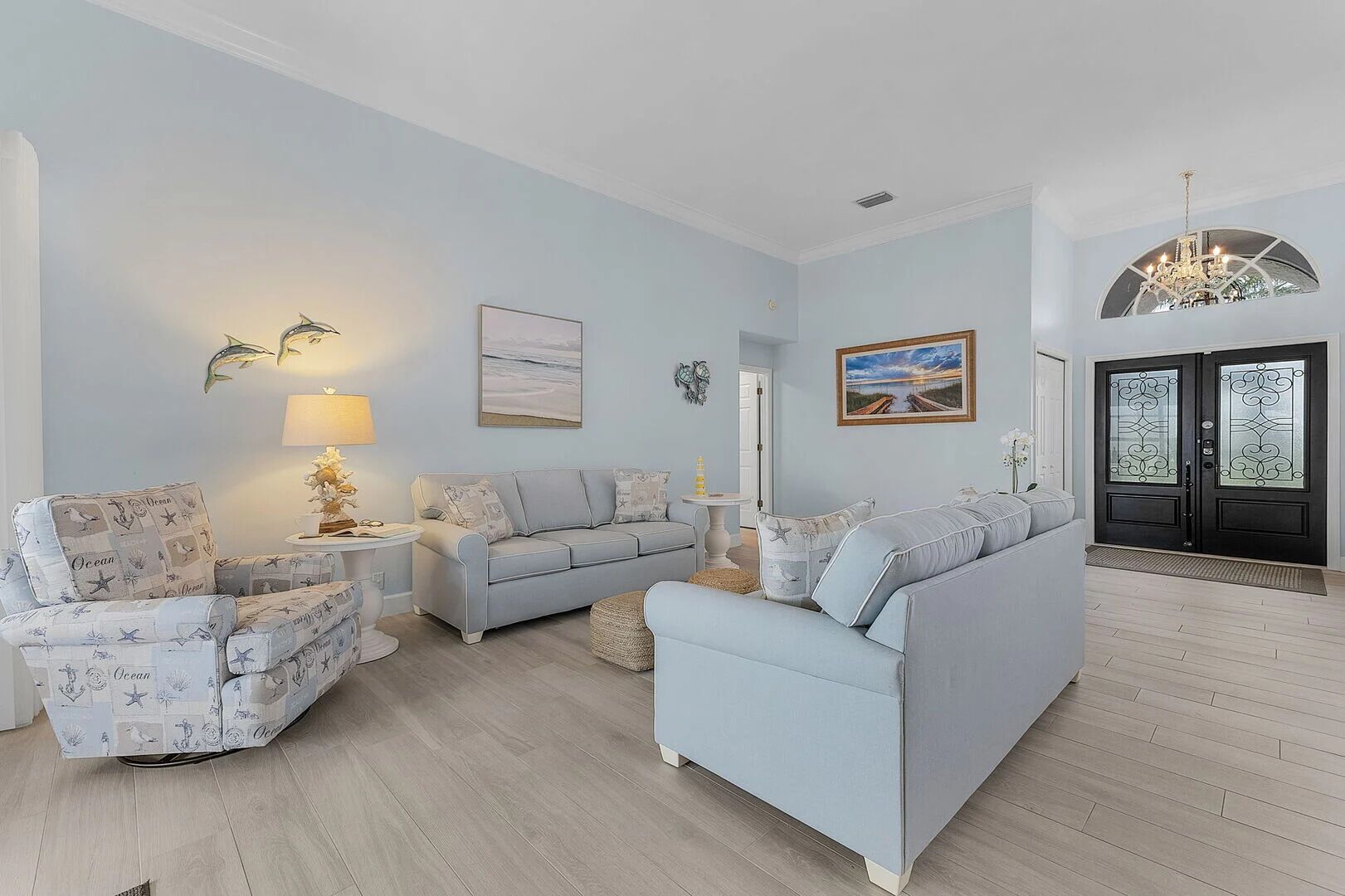 Una sala de estar con un sofá azul claro, un sillón estampado y una pequeña mesa con lámpara. Se observa un cuadro enmarcado en la pared y una puerta con paneles de vidrio decorativo.