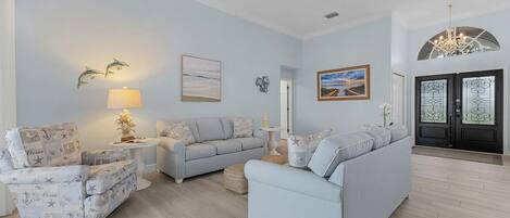 Una sala de estar con un sofá azul claro, un sillón estampado y una pequeña mesa con lámpara. Se observa un cuadro enmarcado en la pared y una puerta con paneles de vidrio decorativo.