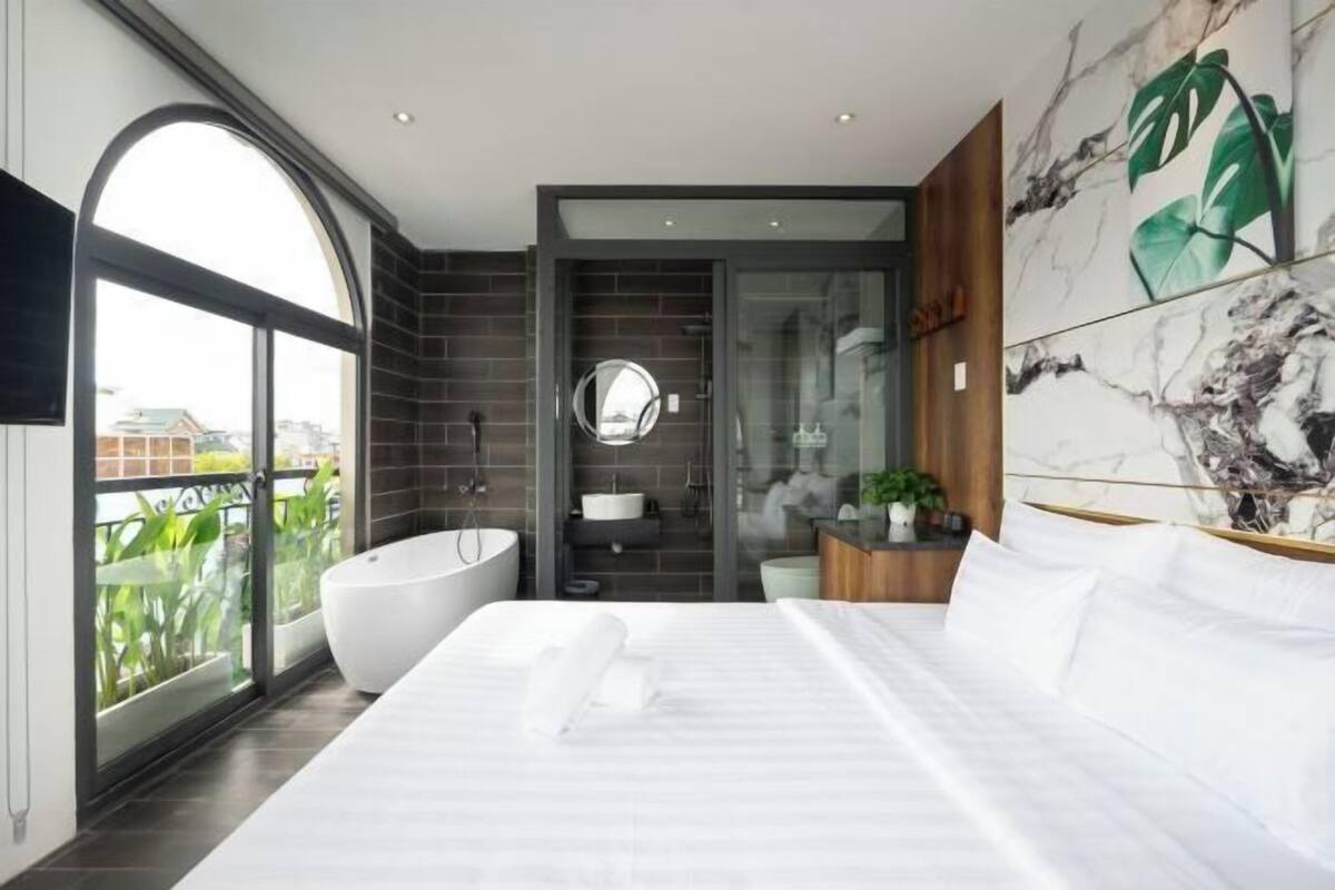 Ein modernes Schlafzimmer mit einem großen Bett, einer freistehenden Badewanne und Blick auf die Stadt durch ein großes Fenster.