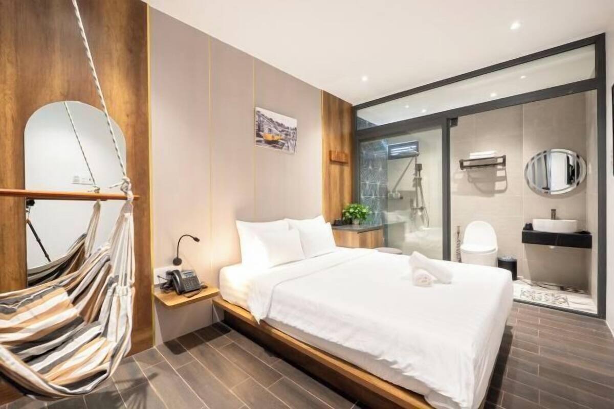 Ein modernes Hotelzimmer mit Bett, Hängematte, Nachttisch mit Telefon und einem Badezimmer mit Glasdusche.