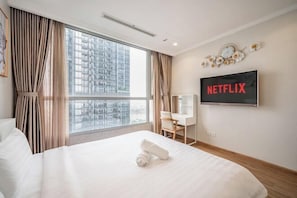 A hotel room with a large bed, a desk, a chair, a TV mounted on the wall, and a window with curtains. - Cozy Haven Apartment - Vinhomes Central Park (Ho Chi Minh City)