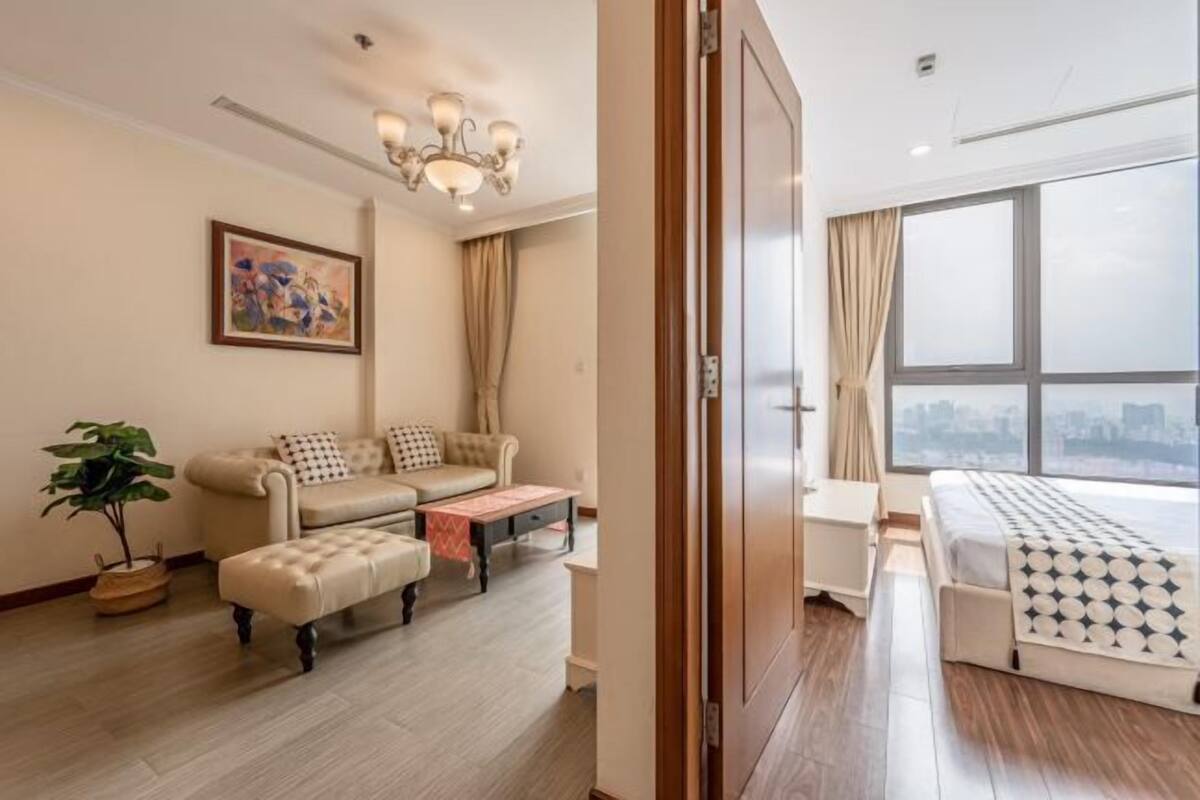 A modern hotel room with a sofa, ottoman, a small table, a bed with a patterned bedspread, a large window, and a city view.