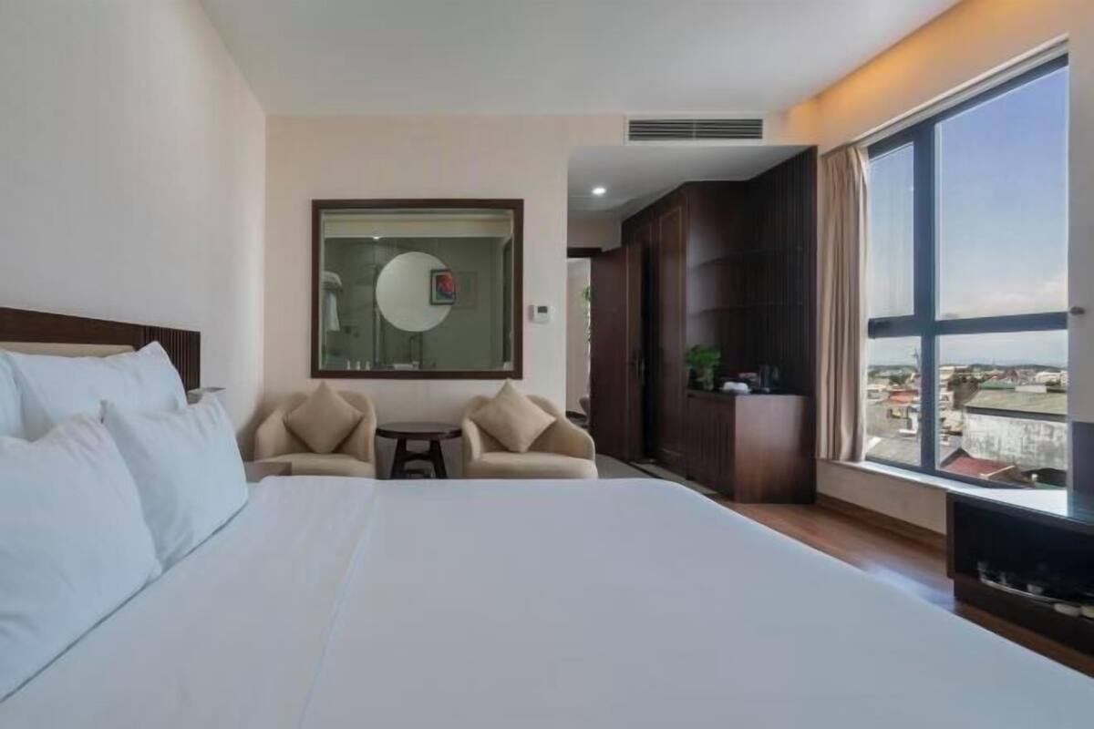 A hotel room with a large bed, a small table, two armchairs, a mirror, and a view of the city through a large window.