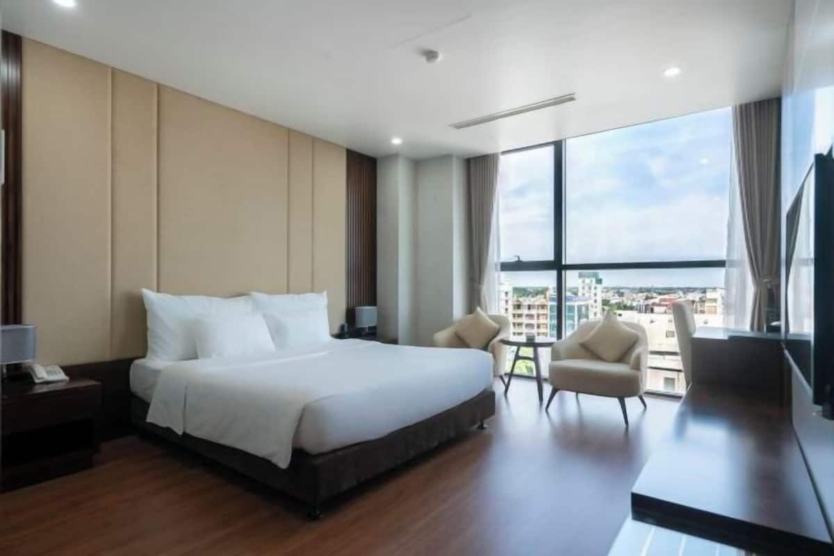A hotel room with a large bed, a desk, two chairs, a small table, and a large window with a city view.