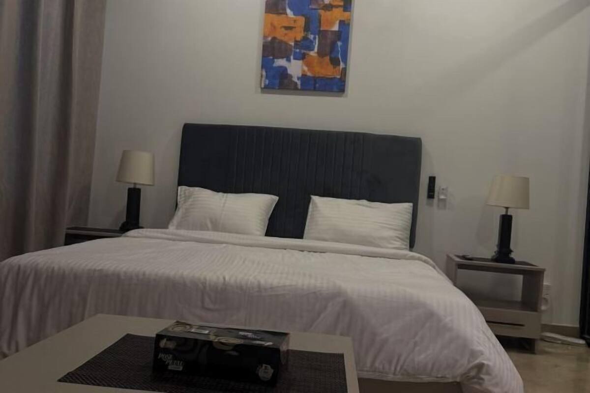 A hotel room with a bed, two bedside tables with lamps, a small table with a box, and a wall painting.