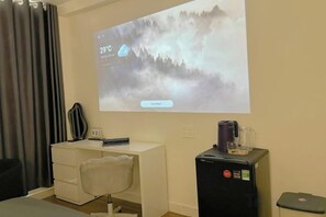 Een hotelkamer met een bed, een bureau met stoel, een scherm waarop een weersvoorspelling staat en een kleine koelkast.