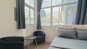 A modern hotel room with a large bed, a small round table, two armchairs, and a view of the city through large windows. - Aesthetic homes Bui Vien Street (Ho Chi Minh City)