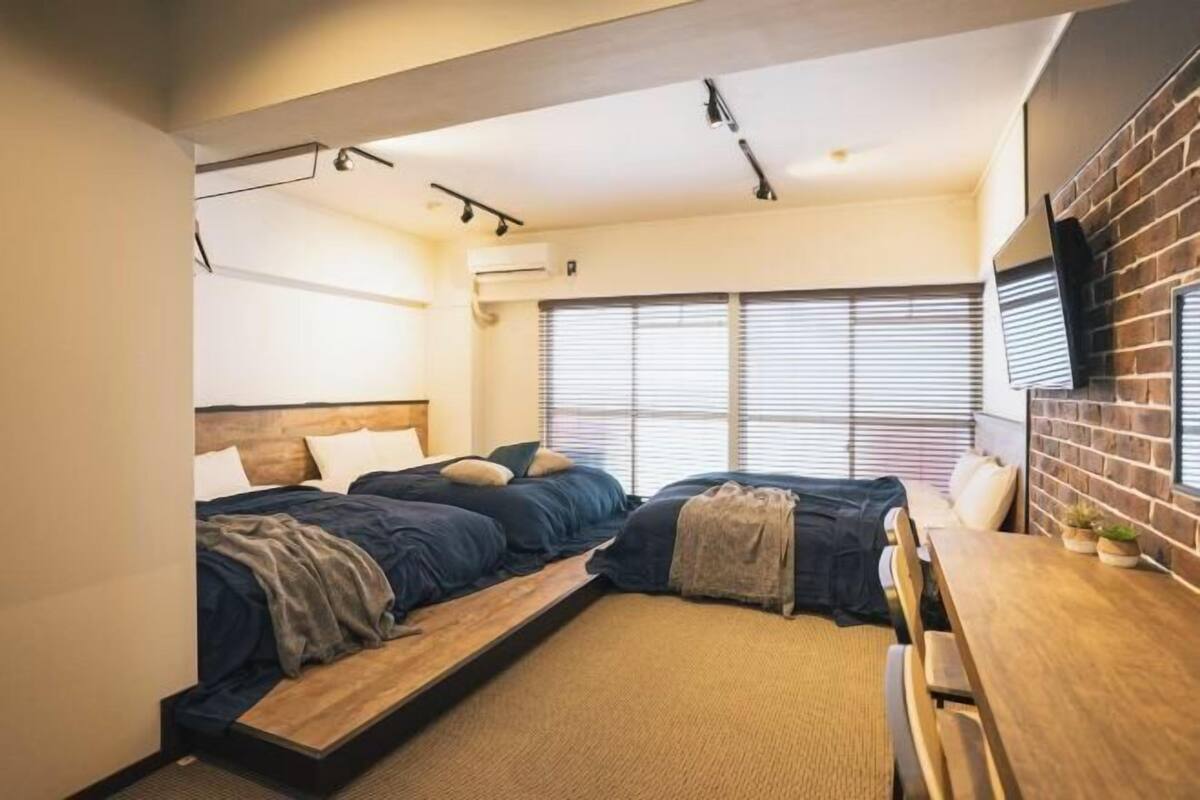 A modern bedroom with two beds, a wooden bench, a TV, and a window with blinds.