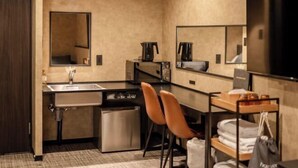 A compact hotel room with a kitchenette, a sink, a small refrigerator, two chairs, a mirror, and a shelf with towels. - Hotel Akasaka Heights (Fukuoka)