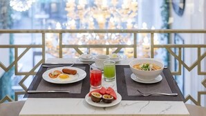 Un tavolo da pranzo con piatti di cibo, un bicchiere di succo e una ciotola di zuppa su un balcone con ringhiera e vista sulla città.