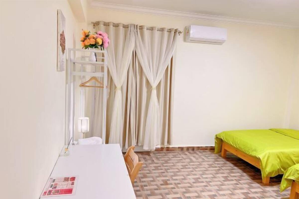 Una stanza arredata con un letto, un tavolo, una sedia, una lampada, uno scaffale con fiori e tende.