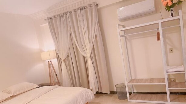 A bedroom with a bed, curtains, a bedside lamp, a white wardrobe, and a small trash bin. - Lemon Tree & Home 2 (New Cairo City)