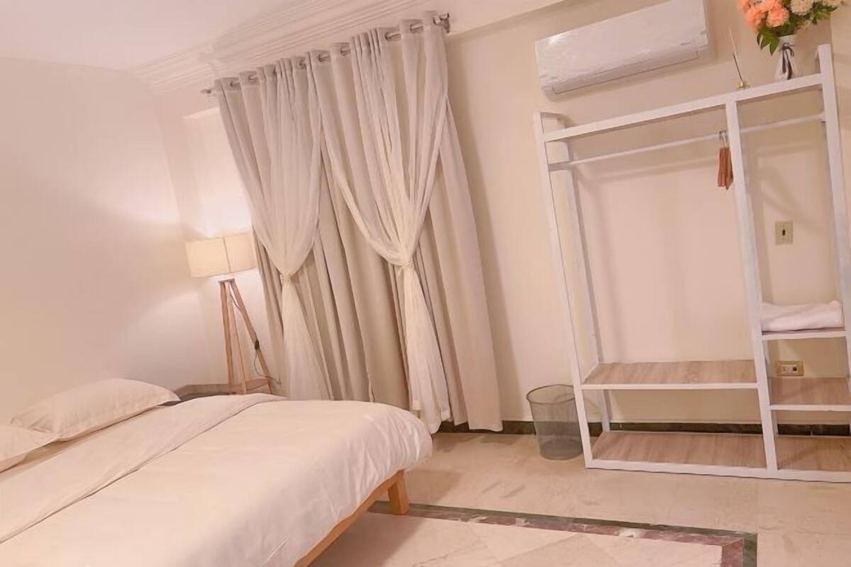 Una camera da letto con un letto, tende, una lampada da comodino, un armadio bianco e un piccolo cestino.