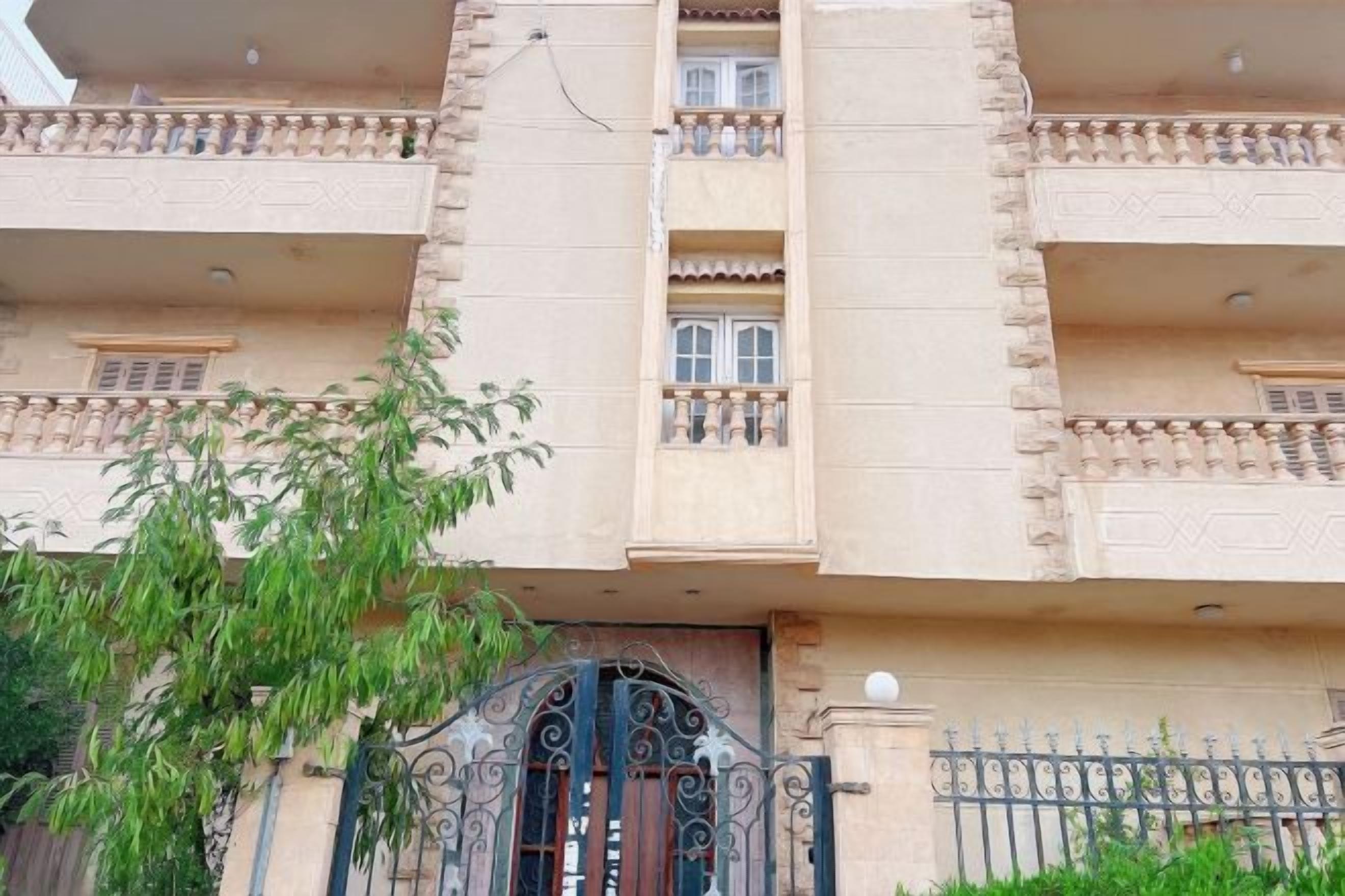 Un edificio de varios pisos con balcones, una entrada protegida por una reja y un árbol en primer plano.