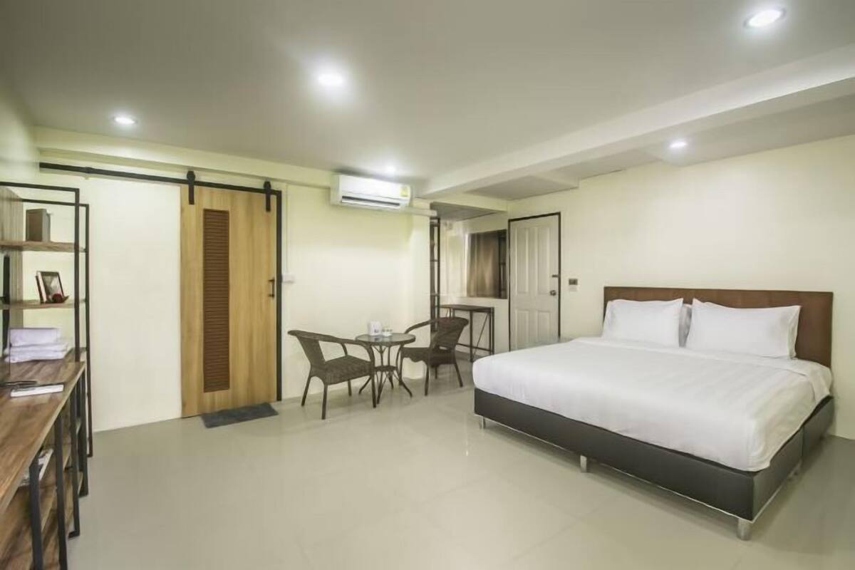 A hotel room with a bed, a desk, a chair, a small table, and an air conditioning unit.