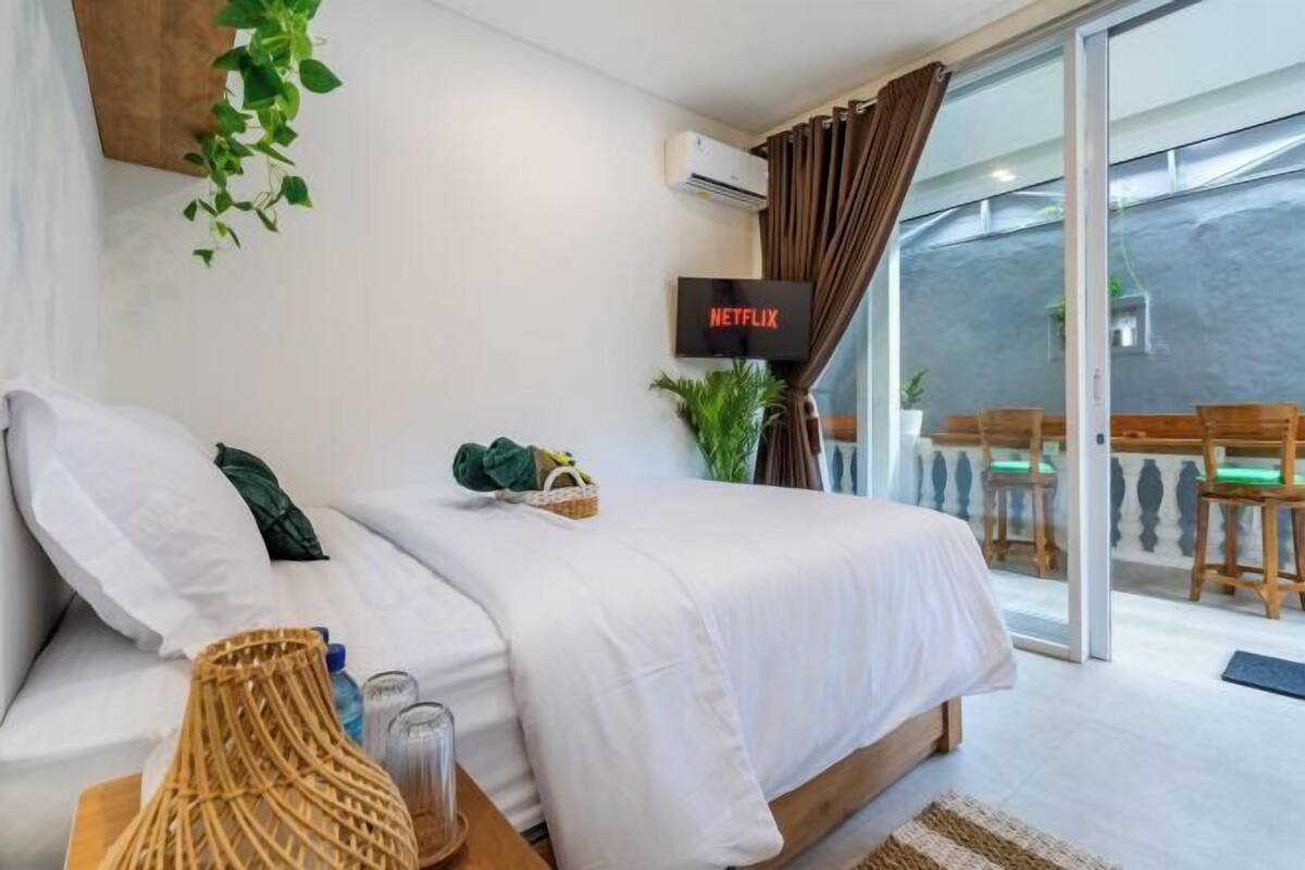 A bedroom with a bed, a TV, a plant, and a balcony with chairs.