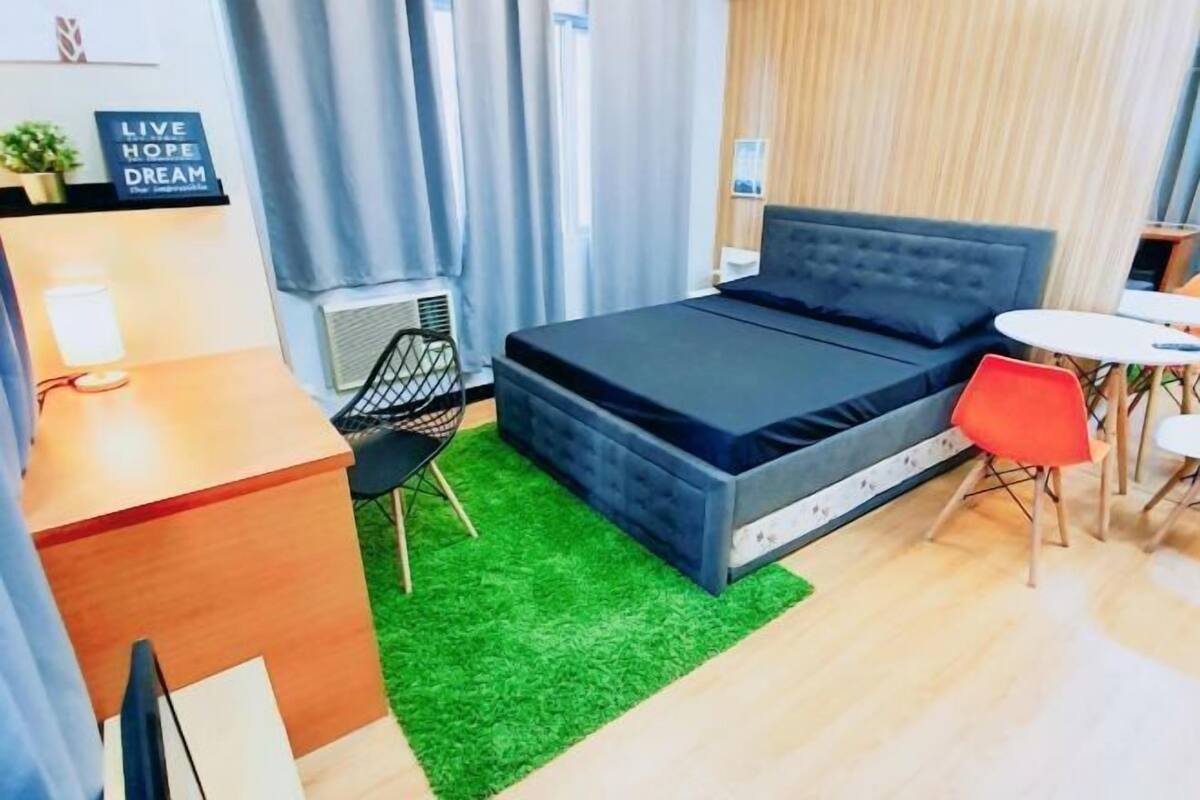 A modern hotel room with a bed, a desk, a chair, a small table, and a green rug.