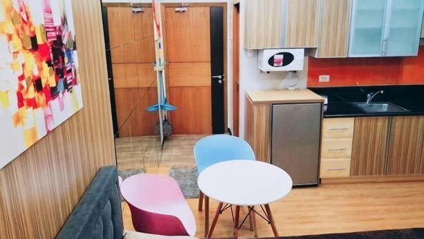 A compact kitchen with a refrigerator, a sink, and a microwave. There are two chairs and a small table. A colorful abstract painting is on the wall. - Maison at the Hill at Morgan Suites Mckinley Hill BGC Taguig (Taguig)