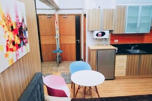 A compact kitchen with a refrigerator, a sink, and a microwave. There are two chairs and a small table. A colorful abstract painting is on the wall. - Maison at the Hill at Morgan Suites Mckinley Hill BGC Taguig (Taguig)