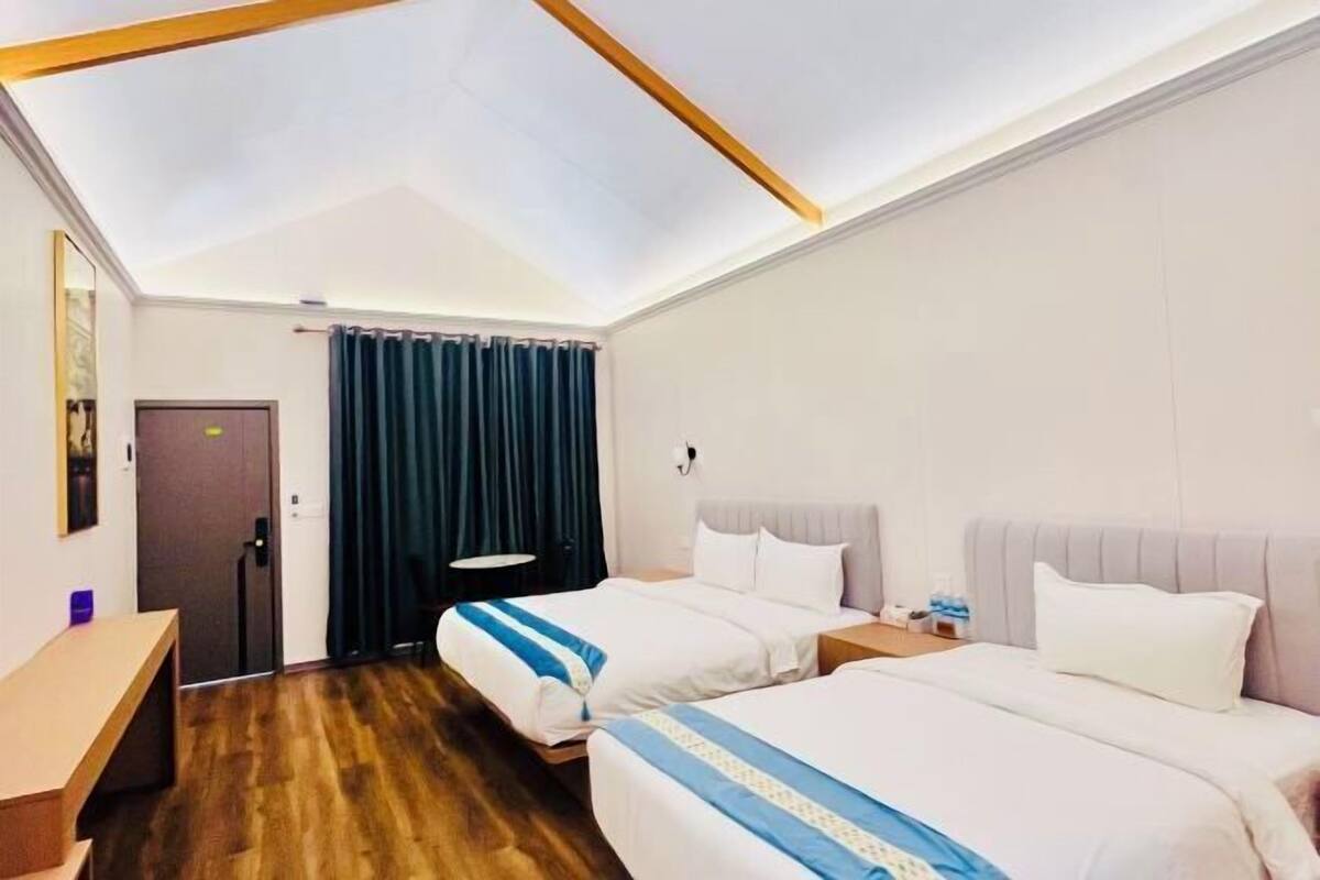 A hotel room with two beds, a desk, a chair, a small table, a nightstand, a lamp, and a window with curtains.