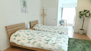 Een tweepersoonsbed met een sprei met bladpatroon, een houten hoofdbord, een groen vloerkleed, een kamerplant en een deur die leidt naar een balkon.