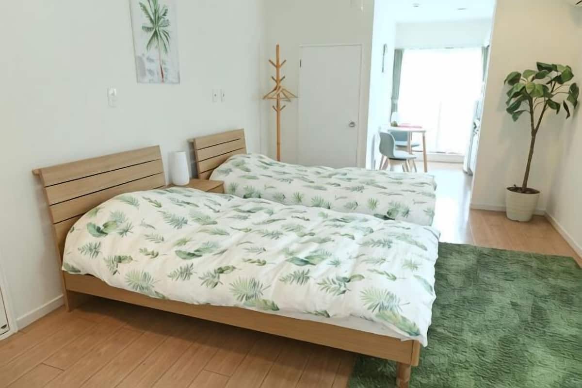 A double bed with a leaf-patterned bedspread, a wooden headboard, a green rug, a potted plant, and a door leading to a balcony.