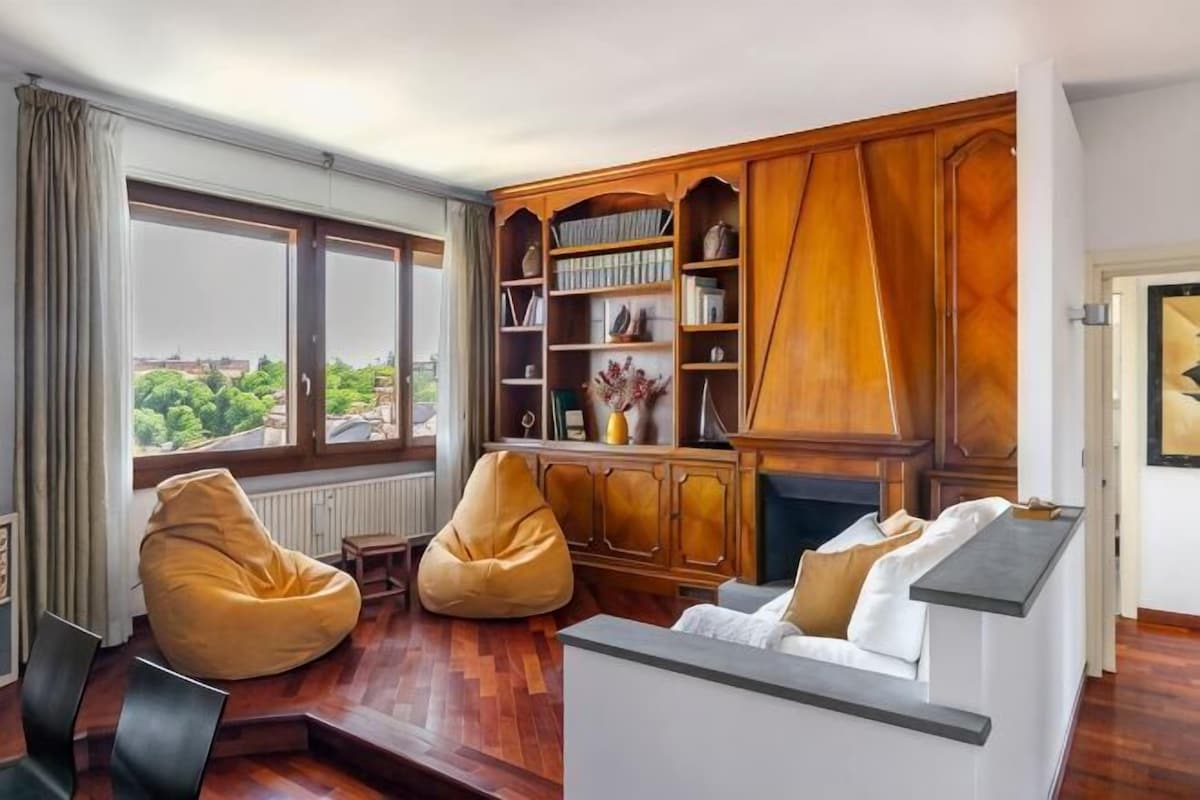 Sala de estar com uma janela grande, estantes de madeira, um sofá branco e duas poltronas de espuma.