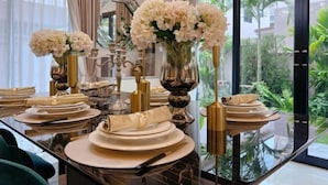Una mesa de comedor con platos blancos, cubiertos dorados y flores blancas en un florero.