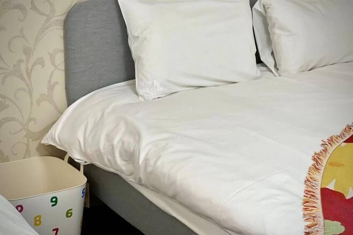 A bed with white bedding and a grey headboard, a white bedside table with a number pattern, and a partial view of a patterned wall.
