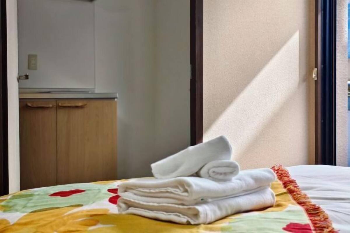 A hotel room with a bed, a stack of folded towels, and a window with natural light.