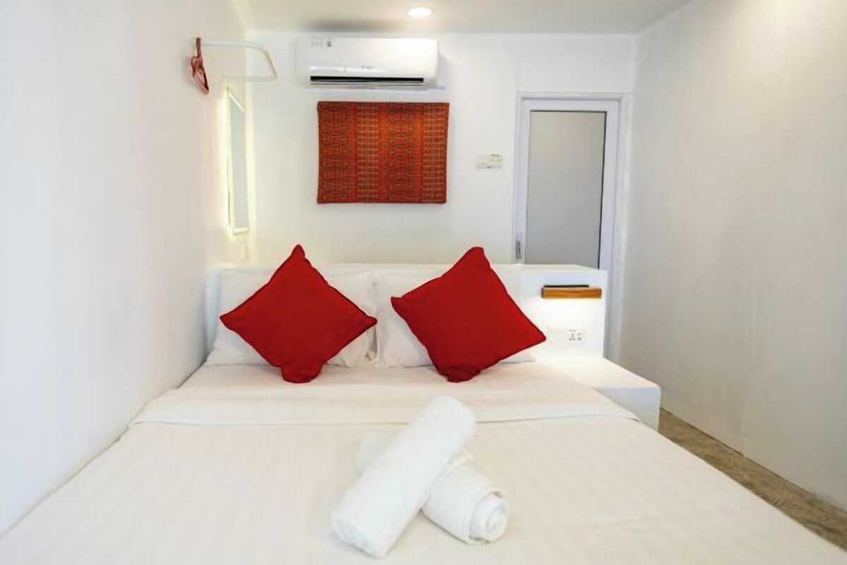 A hotel room with a bed, two red pillows, a rolled white towel, an air conditioning unit, a wall-mounted light, and a door.
