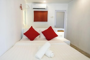 A hotel room with a bed, two red pillows, a rolled white towel, an air conditioning unit, a wall-mounted light, and a door.
