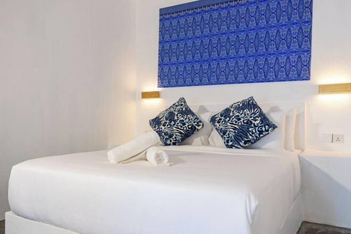 Un lit bien fait, avec une literie blanche et deux coussins à motifs, une décoration murale bleue à motifs au-dessus, et un luminaire fixé au mur sur la droite.