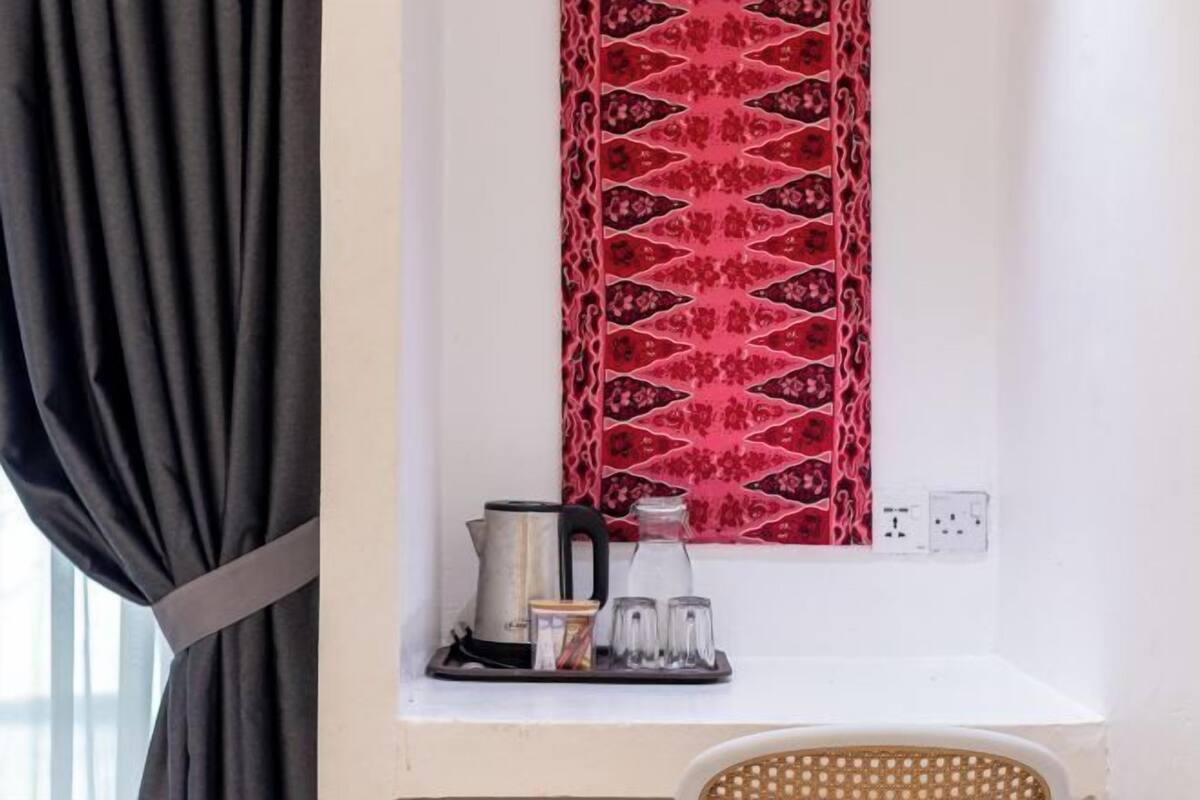 Une fenêtre avec un rideau à motifs, une petite étagère avec une bouilloire, un verre et un plateau, et une prise de courant sur le mur.