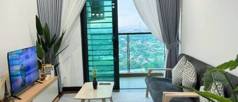 Una sala de estar moderna con televisor, sofá, mesa de centro y vista panorámica de un paisaje urbano a través de un ventanal grande.