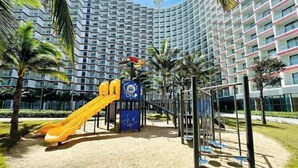 A playground with slides and climbing structures, surrounded by palm trees and a large hotel building in the background. - The KH Arena Cam ranh Nha trang Beachside (Cam Ranh)