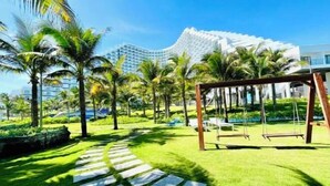 A well-maintained garden with palm trees, a swing set, and a stone pathway leading to a modern building with a unique architectural design. - The KH Arena Cam ranh Nha trang Beachside (Cam Ranh)
