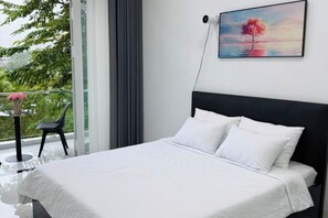 A modern bedroom with a large bed, a view of greenery through a balcony, and a painting on the wall. - Niki Homestay (Hue)
