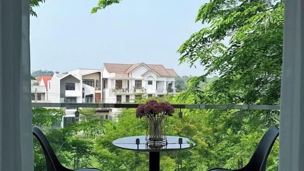 A balcony with two chairs and a table, a vase with flowers, and a view of a multi-story building and greenery. - Niki Homestay (Hue)