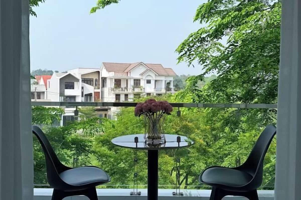 A balcony with two chairs and a table, a vase with flowers, and a view of a multi-story building and greenery.