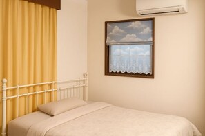 Een bed met een wit metalen frame, een beige dekbed en een raam met uitzicht op wolken en een stadsgezicht.